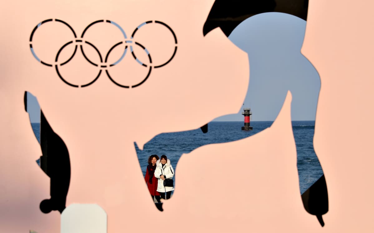 Osittain läpinäkyvä seinä, jonka takaa kaksi naista katsoo kohti kameraa. Seinässä olympiarenkaat ja siluettimainen taitoluistelija, joka on läpinäkyvä.
