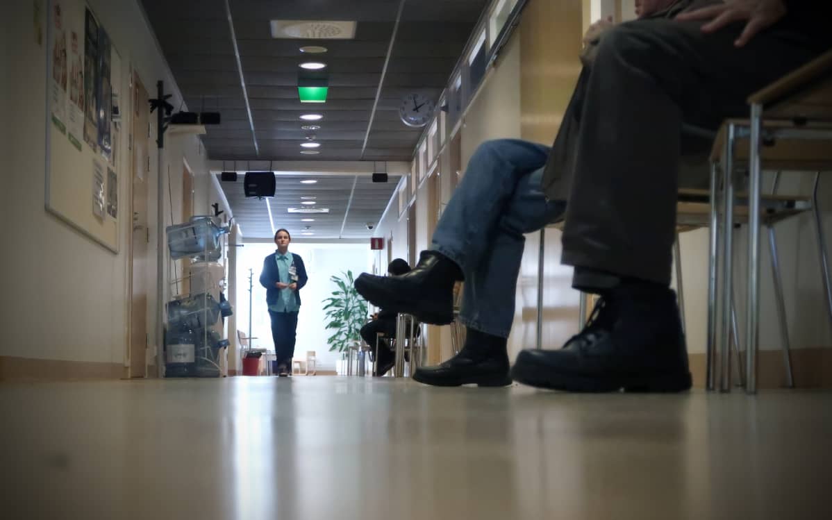 Hoitaja kävelee käytävällä, ihmisten jalkoja etualalla.