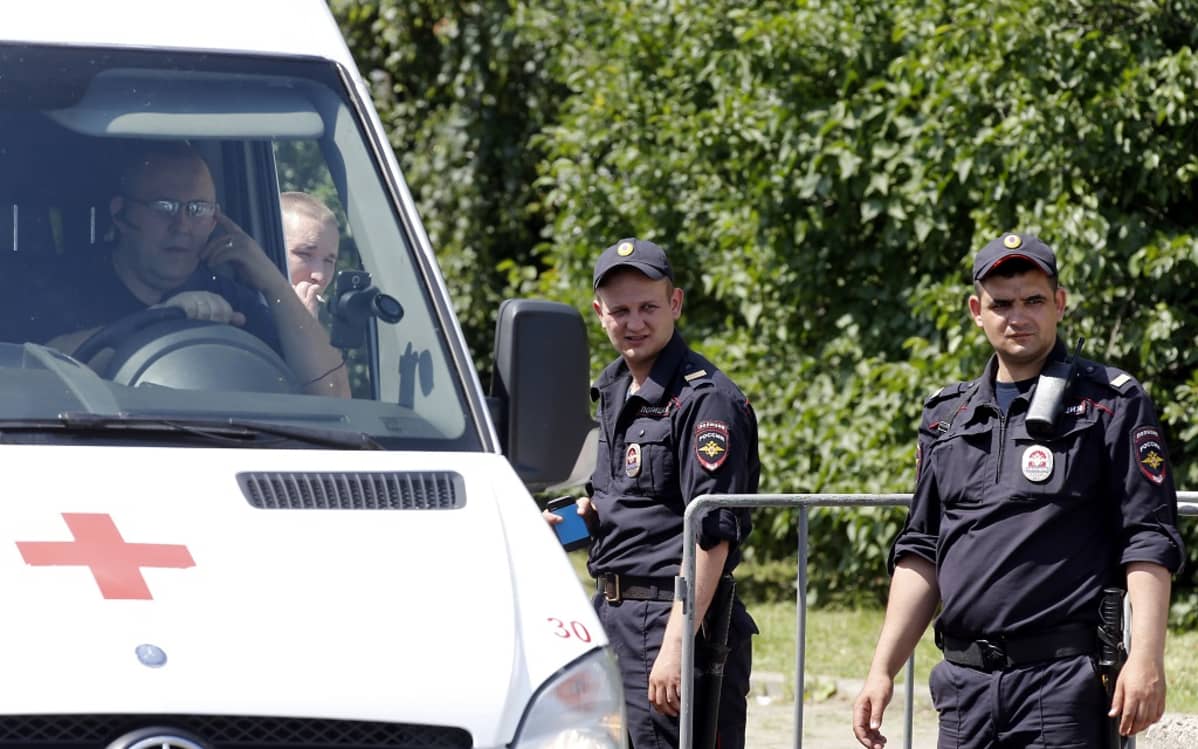 Kaksi univormupukuista miestä seisoo ambulanssin luona. Ambulanssin ratin takana istuu silmälasipäinen mies ja ikkunan läpi näkyy ambulanssin vieressä tupakkaa polttava mies.