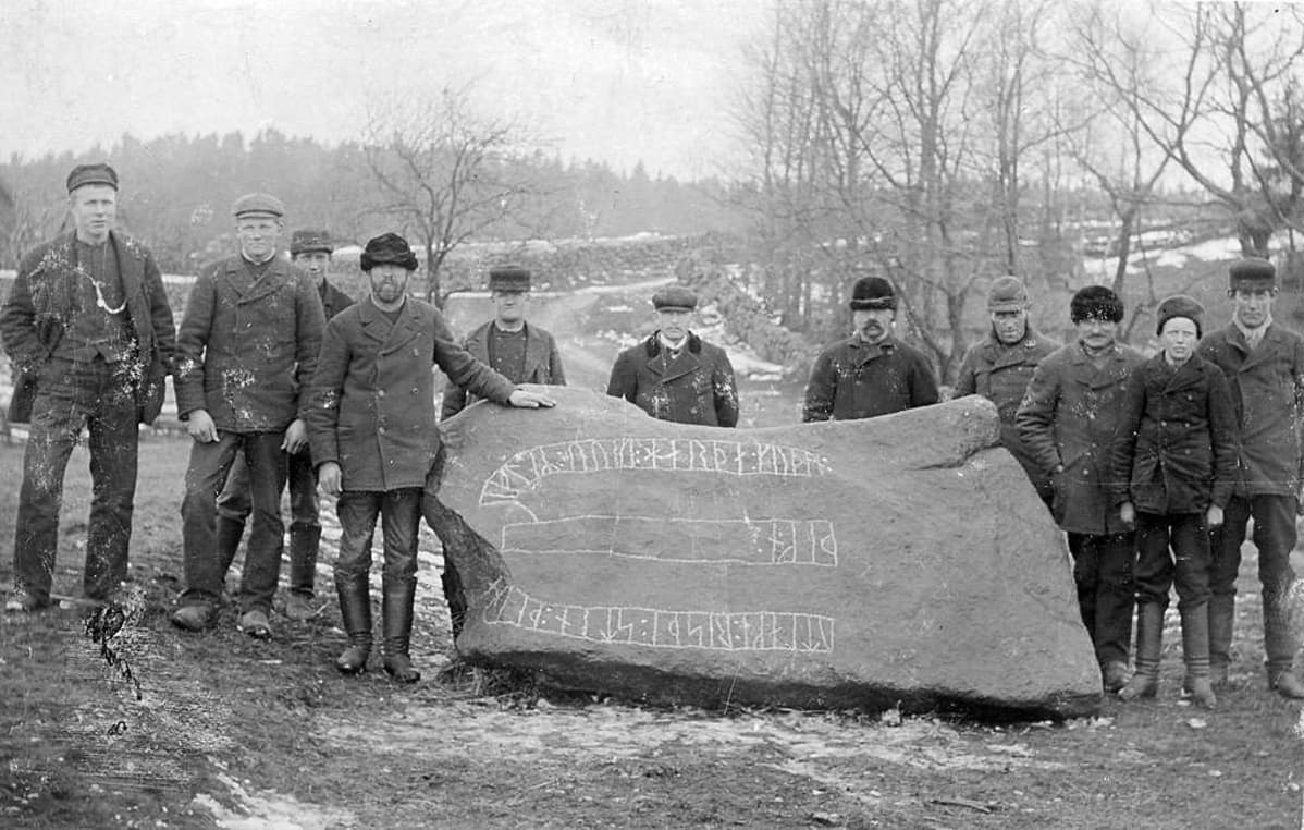 Mustavalkoinen kuba miesjoukosta kyljellään makaavan riimukiven ympärillä.