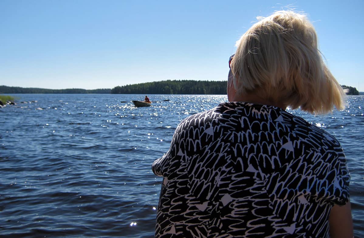 Nainen (selin) istuu järven rannalla, katselee järvellä olevaa soutuvenettä.