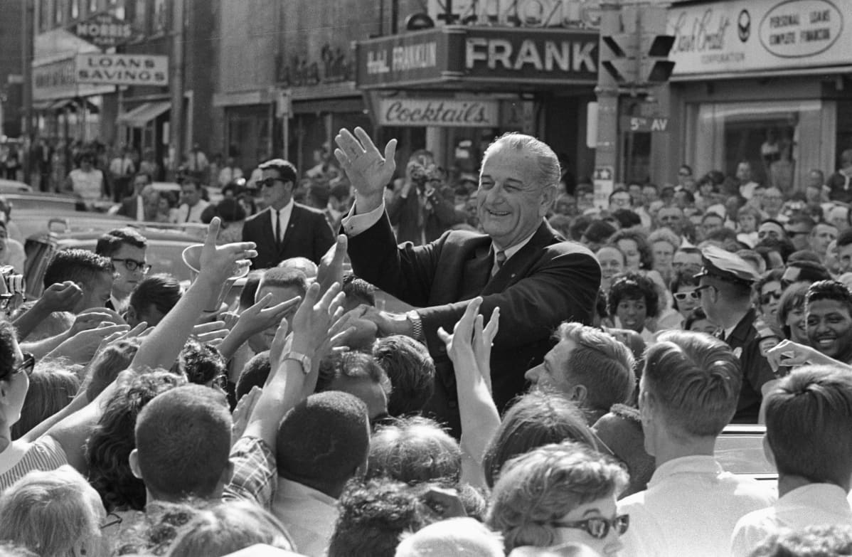 Presidentti Lyndon B. Johnson tervehtii ihmisiä hymyillen väkijoukon keskellä.