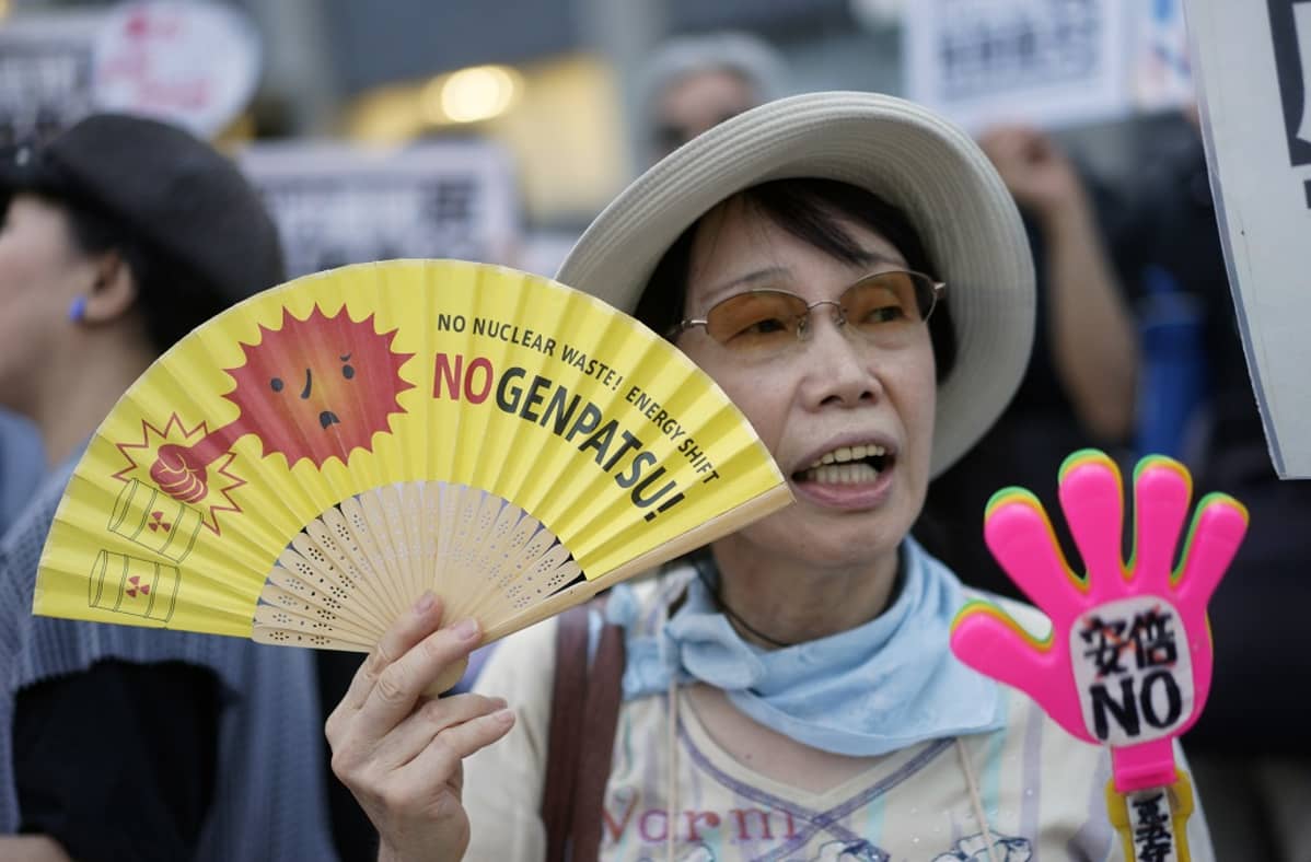 Japanilaisnainen heiluttaa keltaista viuhkaa ja torjuvan käden muotoista kylttiä, joiden teksteissä vastustetaan ydinvoimaa.