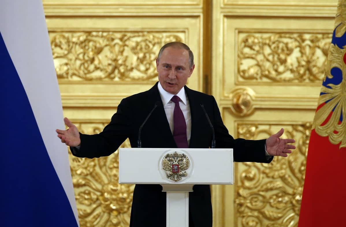 Putin puhuu korokkeen takaa ja levittää juuri käsiään. Putinilla on tumma asu ja viininpunainen kravatti. Taustalla näkyy suuri kullattu ja koristeltu ovi ja kuvan oikeassa ja vasemmassa reunassa Venäjän liput. Korokkeessa on Venäjän vaakunaeläin, kaksipäinen kotka.
