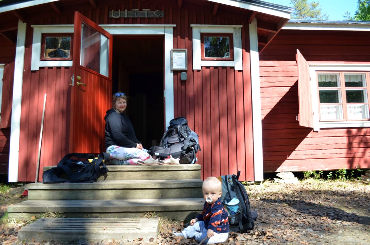 Nainen istuu mökin portailla ja maassa leikkii pieni poika