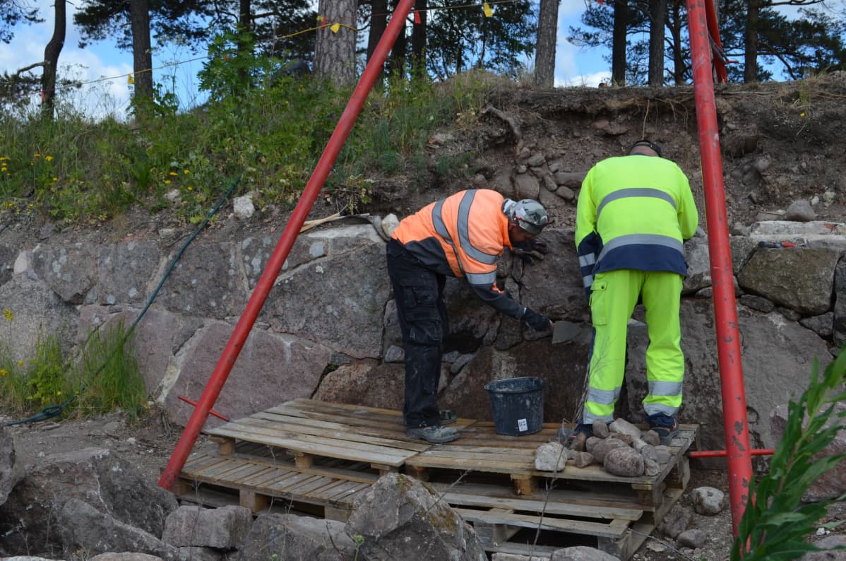 Kaksi miestä työskentelee muurin luona asettaen kiilakiviä isompien muurikivien väliin koloihin.