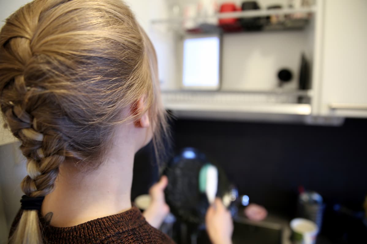 Ponihäntäinen nuori nainen tiskaa etualalla ja mobiililaite on asetettu eteen tiskikaappiin. Näytöltä nainen lukee kirjaa.