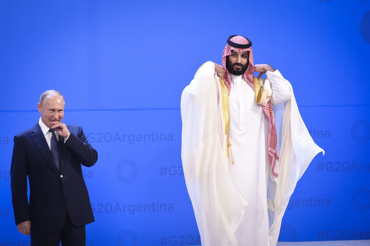 Vladimir Putin hymyilee oikein maittavasti etusormi huulien edessä vekkuli ilme kasvoillaan ja hieman korkeammalla oikella vieressä seisoo Saudi-Arabian kruununprinssi Mohammed bin Salman oikoen valkoista jalabiaan katsoen kohti Putinia.