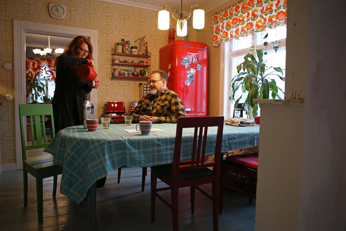 Mies istuu ruokapöydän ääressä ja nainen kaataa kahvia.