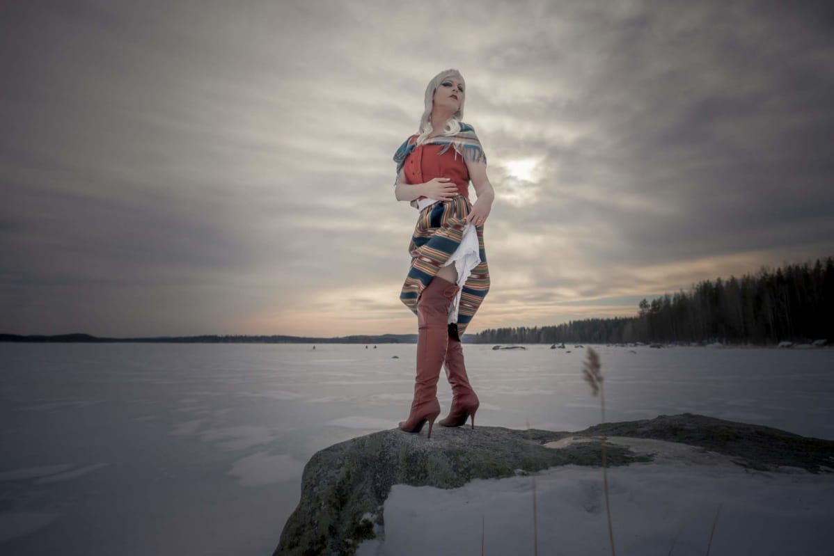 Drag-artisti poseeraa kalliolla kansallispuvussa saapikkaat jalassa ja nostaa hameensa helmaa