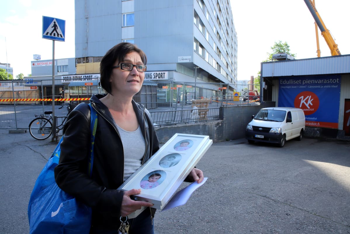 Nainen kantaa käsissään kahta kehystä, joissa valokuvia. Naisen takana kerrostalo