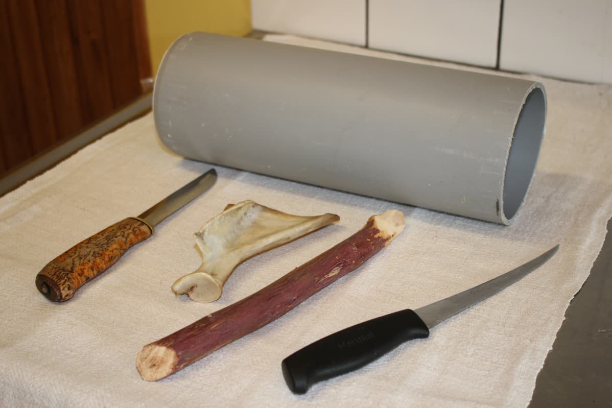 Pöydällä liinan päällä puukko, lusa, keppi, veitsi ja putkenpätkä.
