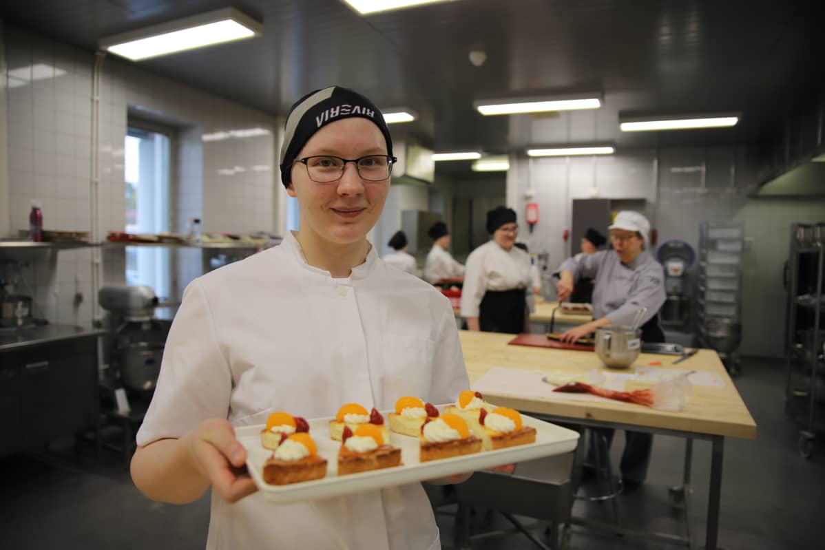 Nuori leipurityttö kantaa tarjottimella värikkäitä leivoksia.