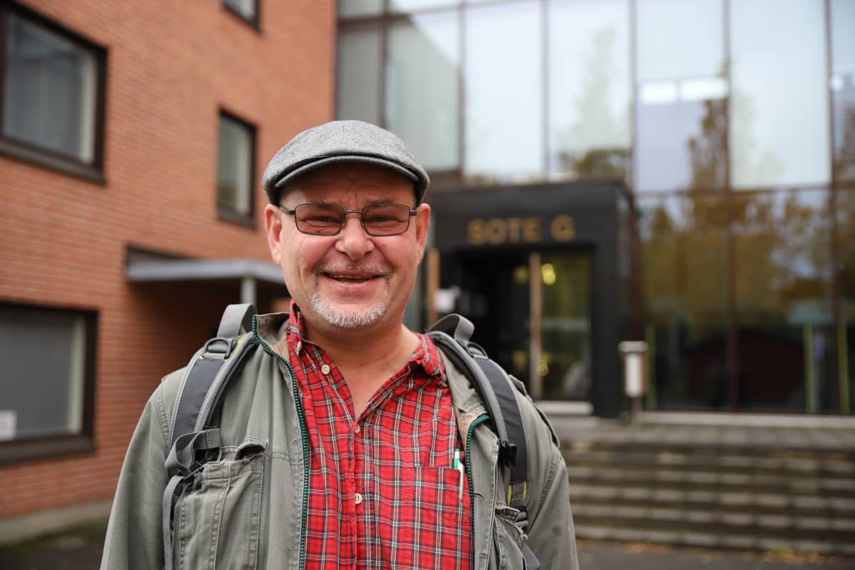 Lippalakkipäinen keski-ikäinen mies hymyilee koulun pihalla.
