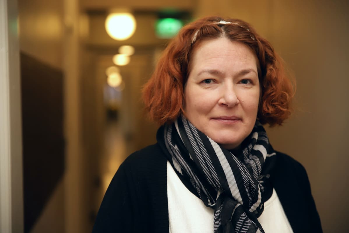 Nainen seisoo käytävässä ja katsoo suoraan kameraan.