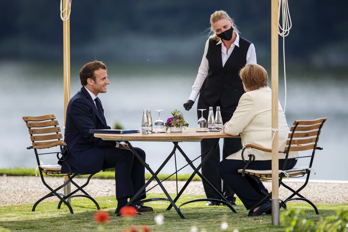 Kuvassa ovat Emmanuel Macron, Angela Merkel ja mustaa hengityssuojainta käyttävä tarjoilija.