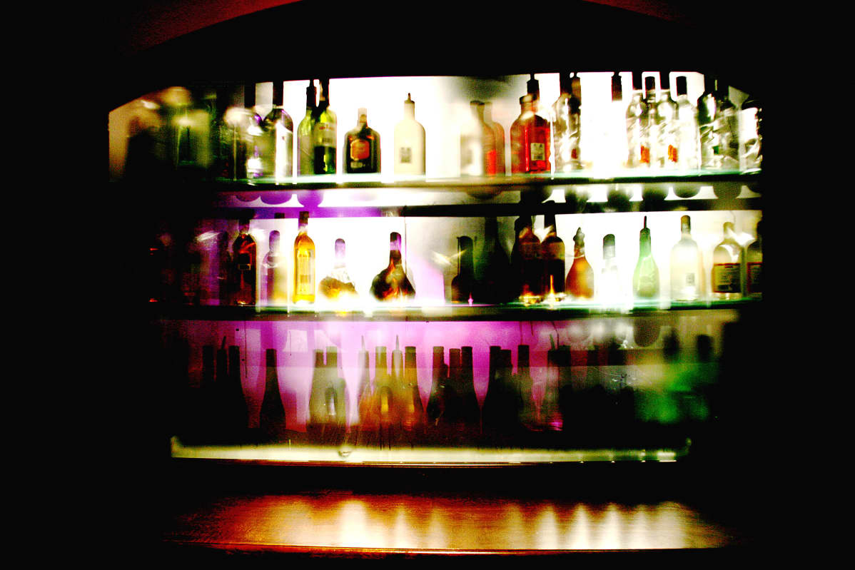 Alkoholipulloja baarin kaapissa rivissä, taustalta tulee valoa pullojen läpi.