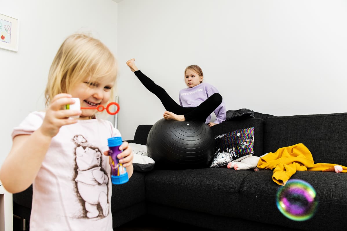 Sofia puhaltaa saippukuplia, Alina temppuilee sohvalla.
