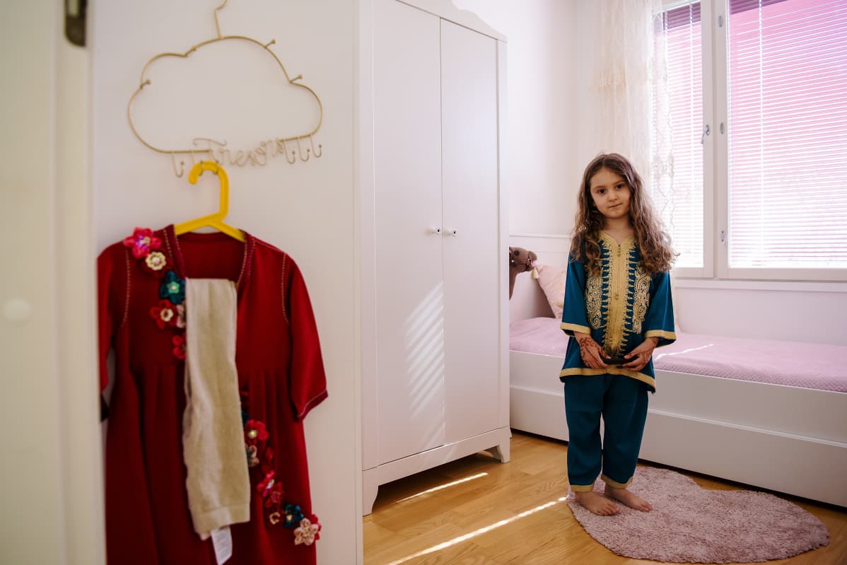 Liliya poseeraa huoneessaan, vasemmalla odottavat huomisen juhlapäivän vaatteet.