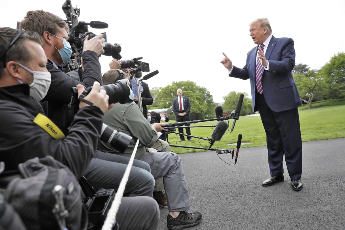 Donald Trump puhuu toimittajajoukolle. Trumpilla on tumma puku ja raidallinen kravatti. Hänen eteensä työnnetään mikrofoneja, Kuvaajilla on kasvomaskit. Kauempana taustalla seisoo turvamies.