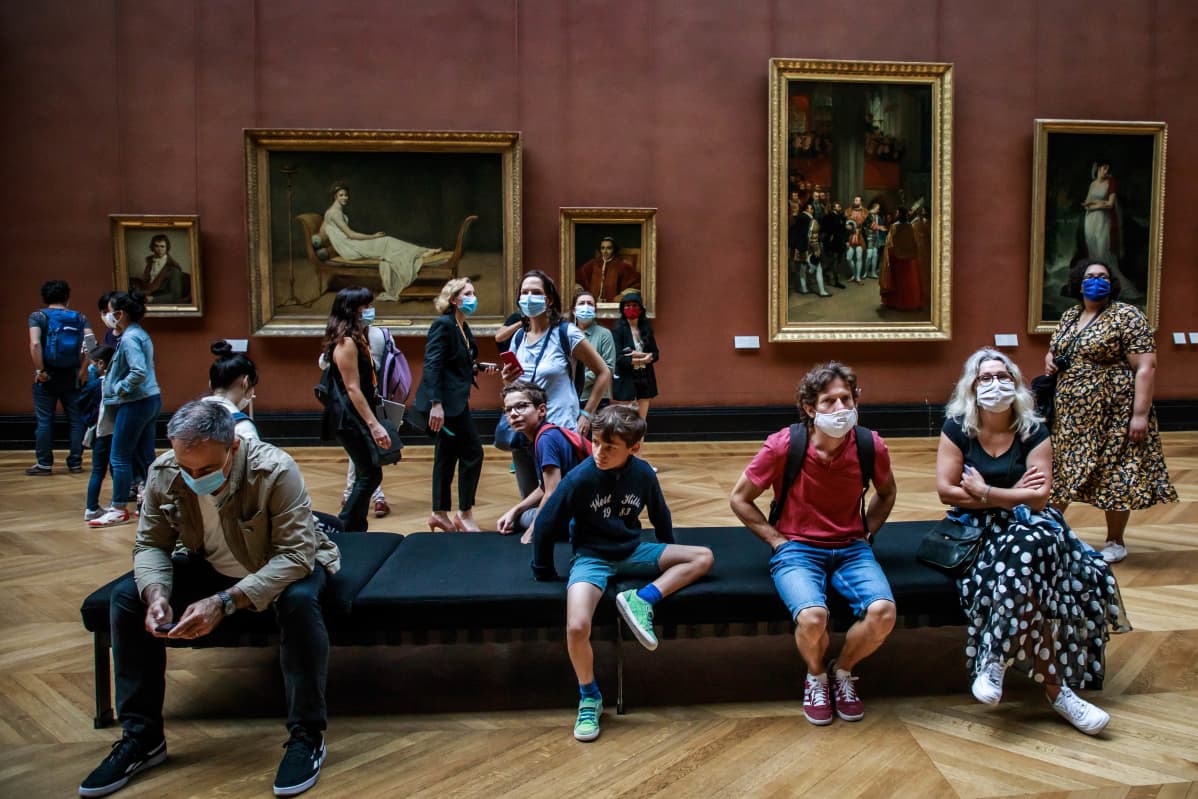 Turisteja maskit kasvoillaan istumassa ja seisomassa museon salissa.