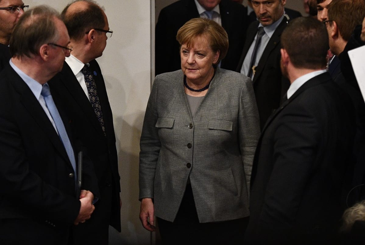 Vakavailmeinen Merkel tulossa huoneeseen, ympärillä pukuihin pukeutuneet miehet ovat kääntyneet katsomaan häntä.