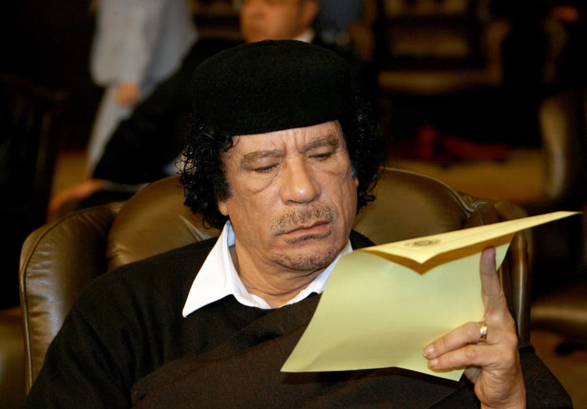 Gaddafi istuu nojatuolissa ja lukee paperia. Kyseessä on Arabiliigan kokous Damaskoksessa 2008, ja taustalla näkyy muita kokoukseen osallistujia.