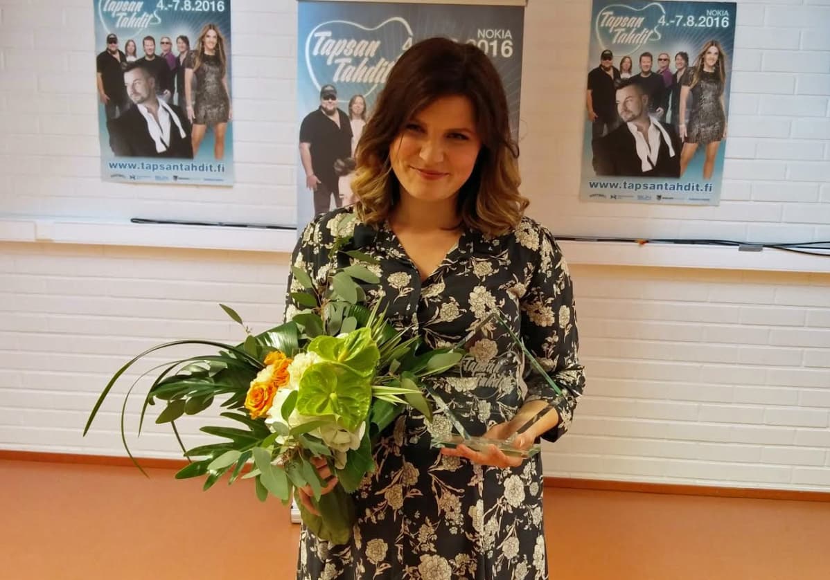 Suvi Teräsniska poseeraa kuvaajalle kukkakimppu ja lasinen Tahti-pysti kädessään.