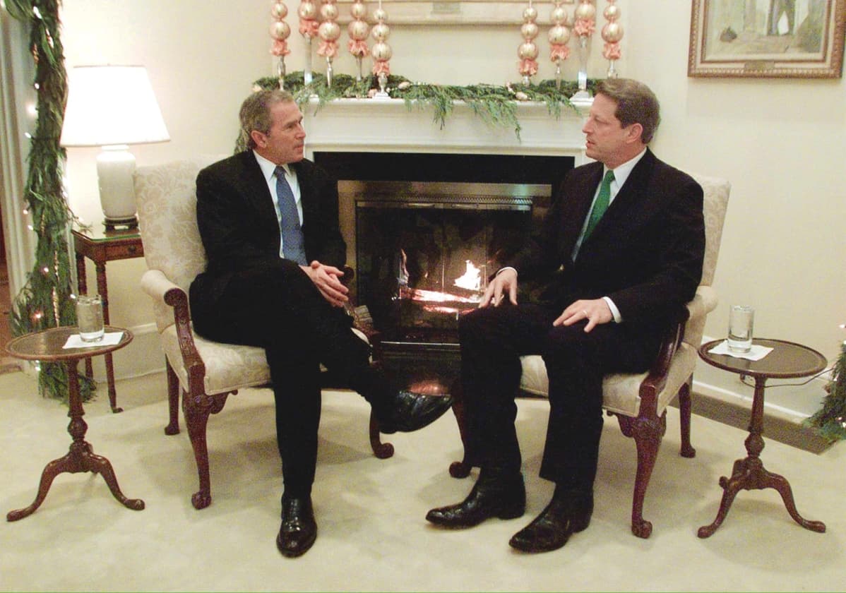 George W. Bush ja Al Gore istuvat tuoleilla. Taustalla takkatuli ja joulukoristeita.