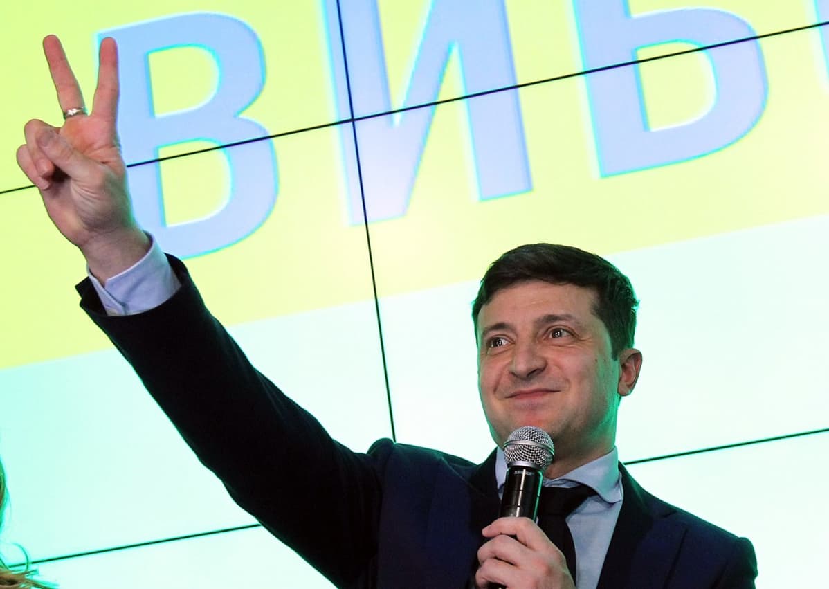 Zelenskyi tekee sormillaan voitonmerkin ja pitelee toisessa kädessään mikrofonia.