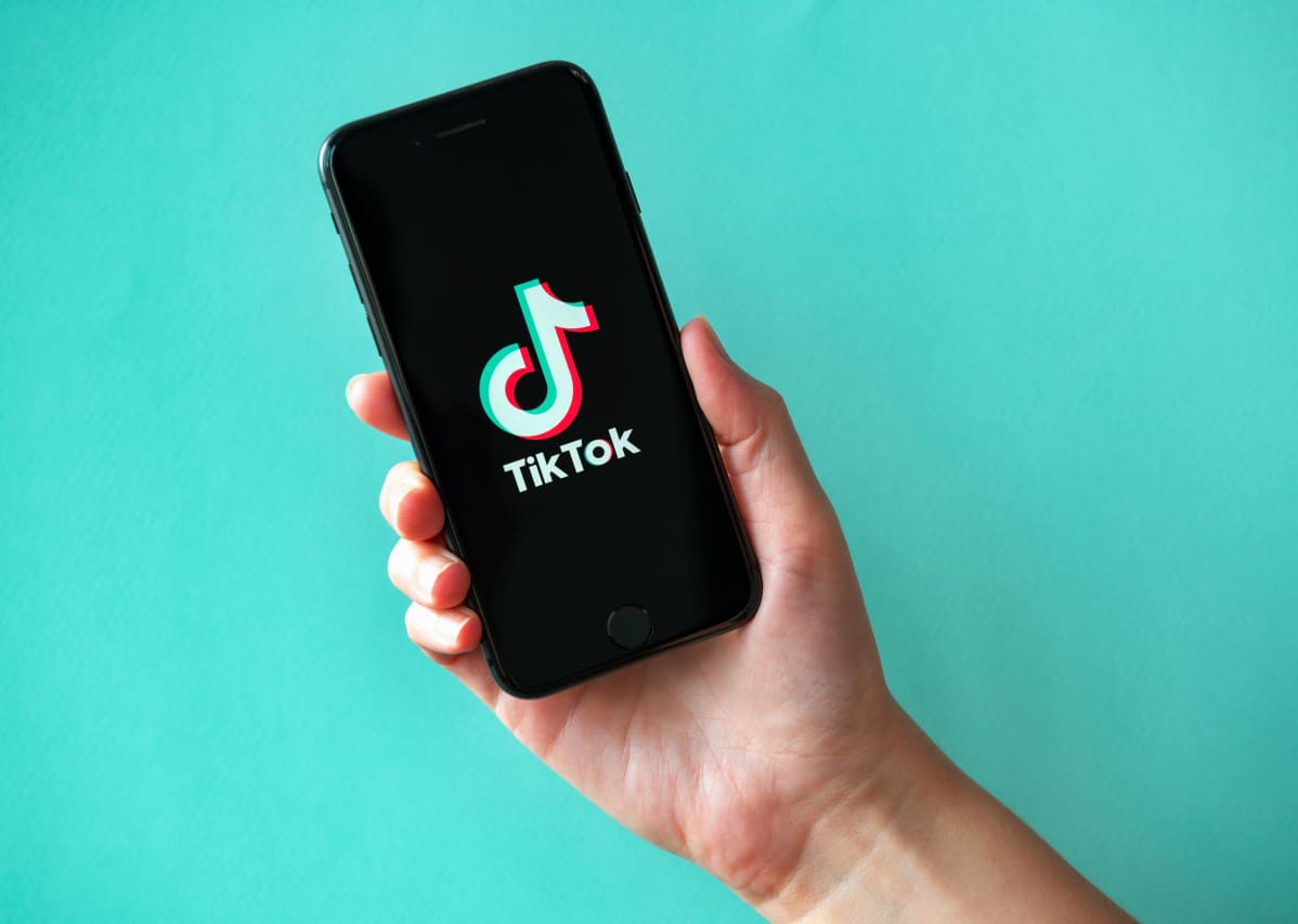 En person håller i en telefon där en logo syns. Det står tiktok. I bakgrunden syns en mintgrön vägg.