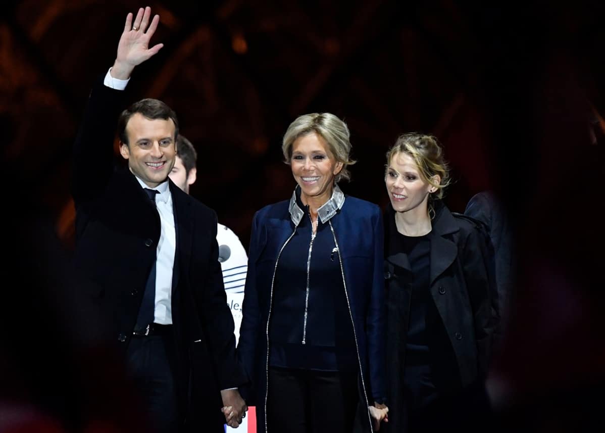 Tummaan pukuun pukeutunut hymyilevä Emmanuel Macron vilkuttaa, vieressään kaksi vaaleaa naista.