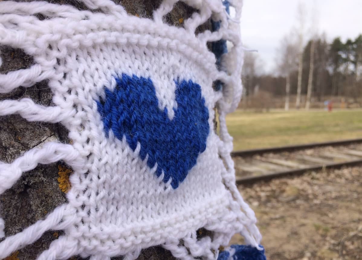 Neulegraffitityö, jossa on sininen sydän valkealla pohjalla. Työ on kiinnitetty puun runkoon ja taustalla näkyy puisto.