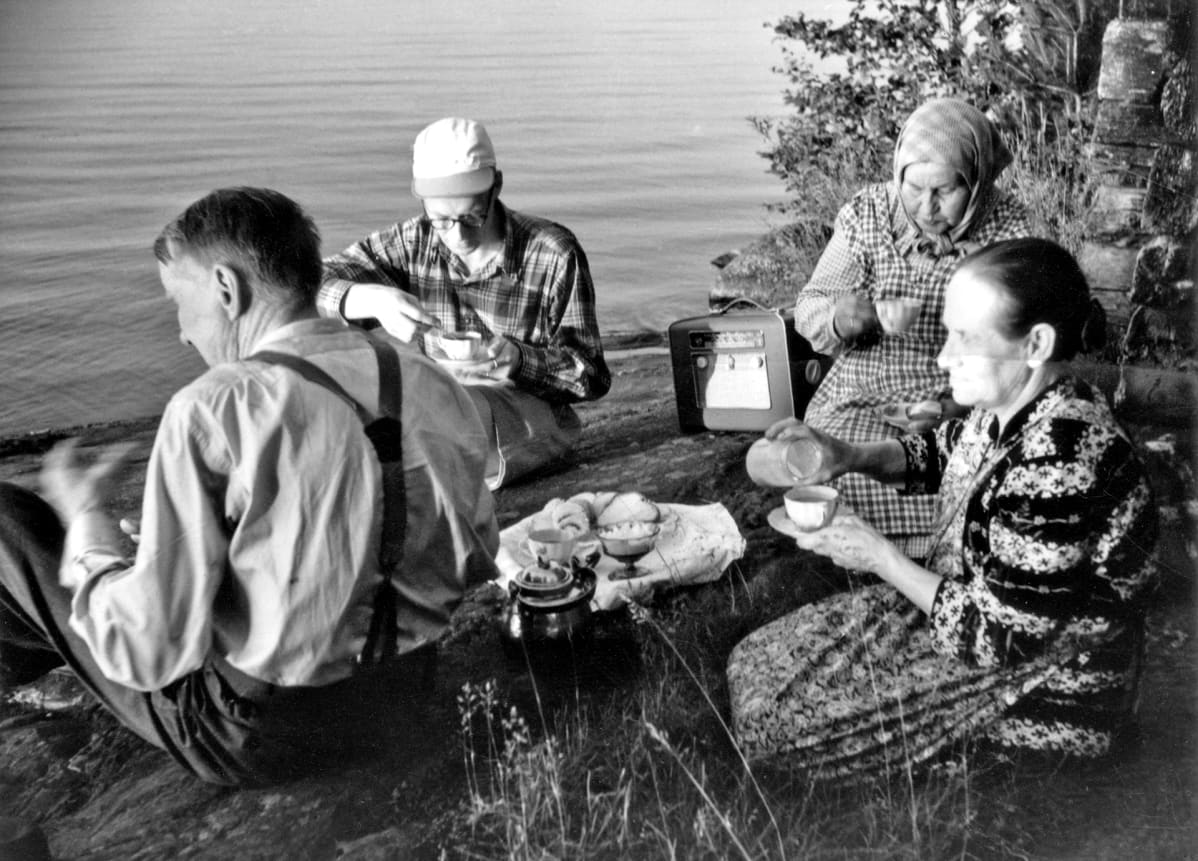 Mustavalkoinen kuva. Seurue juomassa kahvia rannalla, matkaradio mukana kalliolla. Kaksi vanhaa naista, nuori mies ja vanha mies istuvat rantakalliolla ja kuuntelevat radiota.