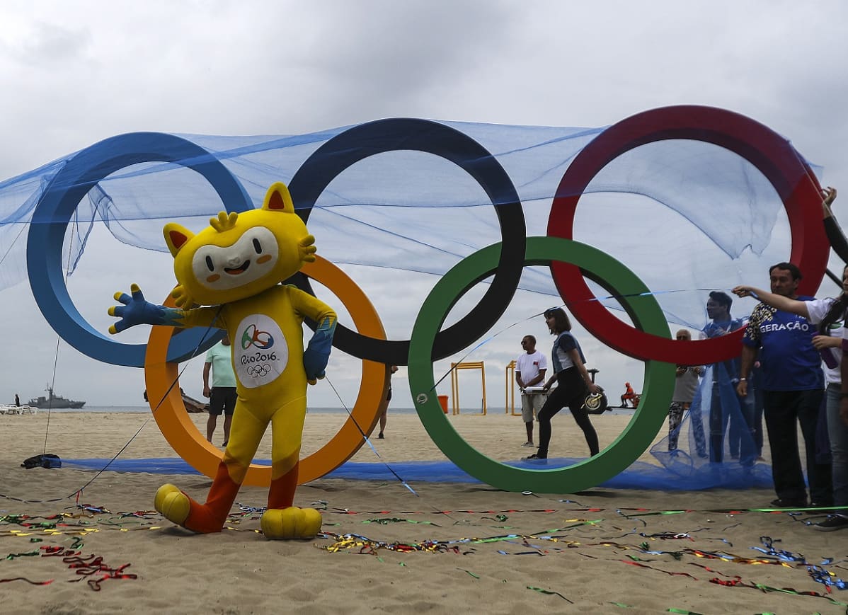 Keltainen, kissalta näyttävä olympiamaskotti olympiarenkaiden edessä, hiekkarannalla.