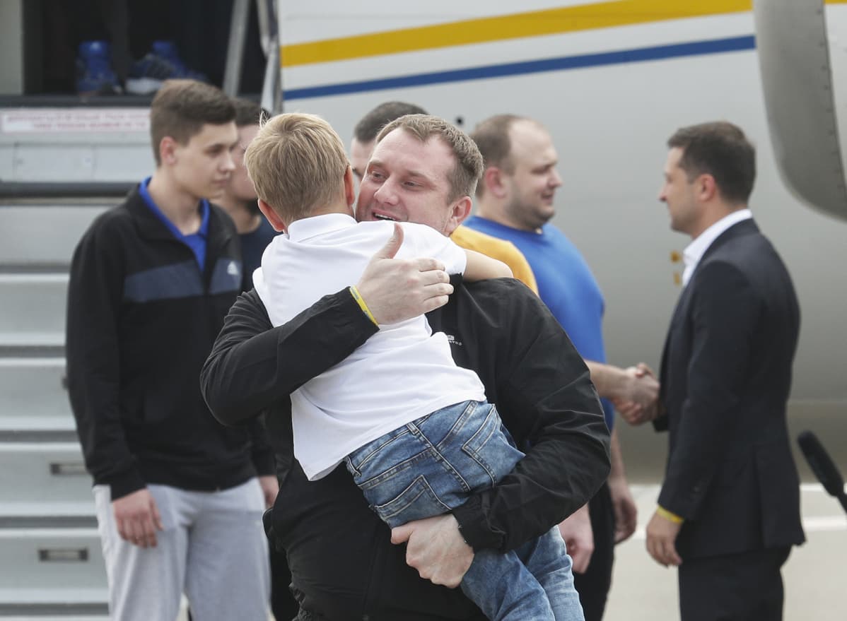 Vapautettu ukrainalainen merimies halaa poikaansa samalla kun Ukrainan presidentti Volomydyr Zelenski kättelee muita vapautettuja vankeja. Taustalla näkyy vankeja tuonut lentokone, jonka kyljessä on Ukrainan lipun väriset raidat.