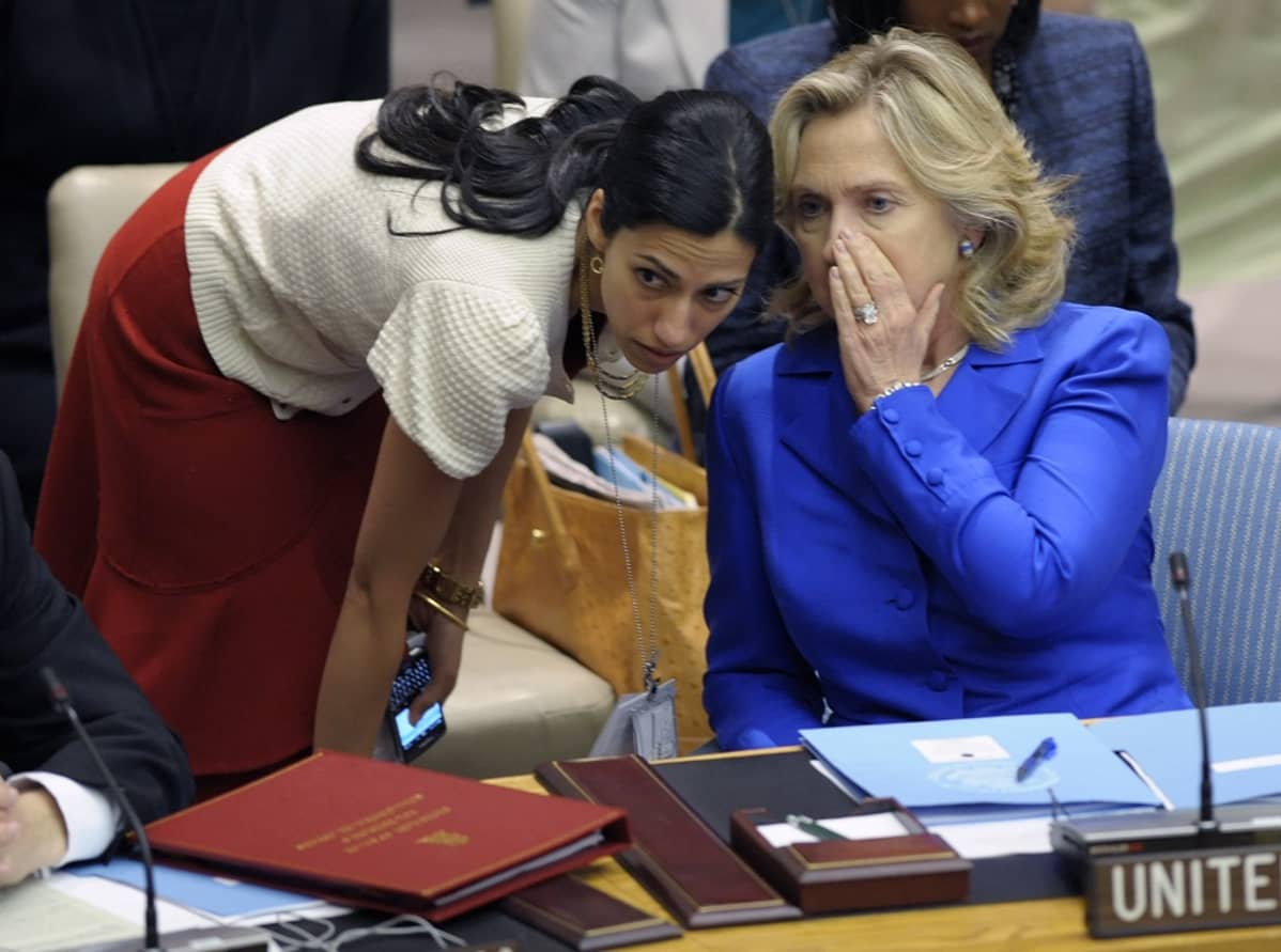Siniseen jakkuun pukeutunut Hillary Clinton kuiskaa valkoiseen neuleeseen ja punaiseen hameeseen pukeutuneelle Huma Abedinille.