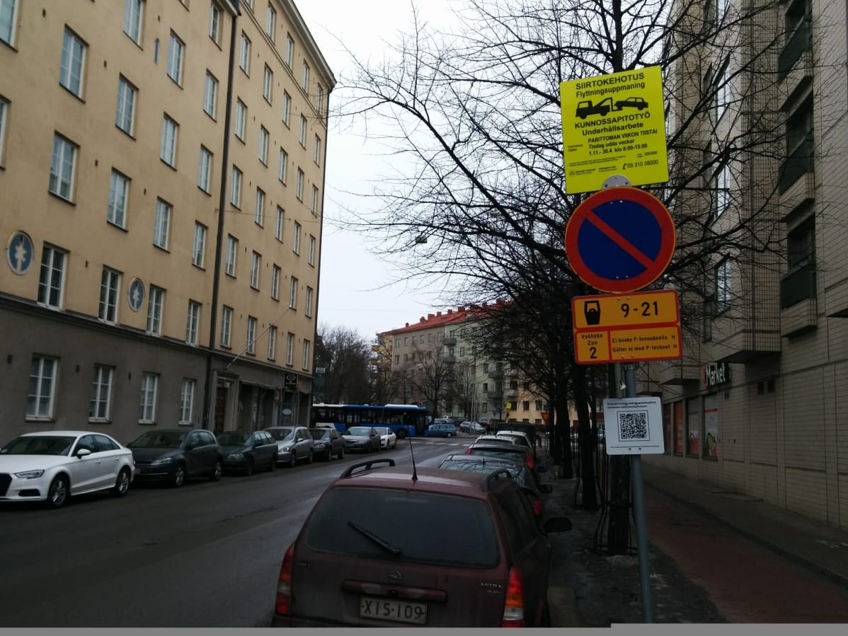 Näkymä kadulta Taka-Töölöstä. Etualalla liikennemerkki, jossa on siirtokehotus. Henkilöautoja parkkeerattu molemmin puolin kerrostalojen reunustamaa katua.