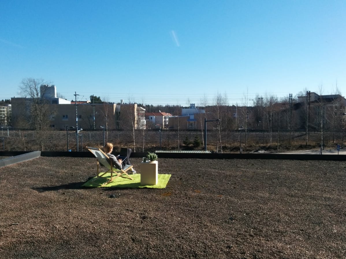 Opiskelija istuu rakennuksen tasakattoisella katolla aurinkotuolissa kannettava tietokone sylissä. Taustalla näkyy junarata ja kerrostaloja.