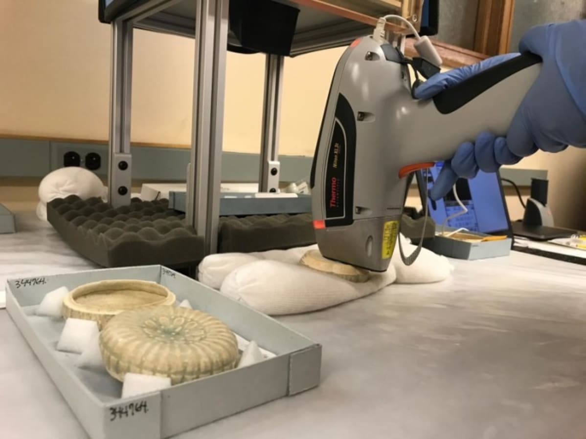 Keramiikkaesineitä laboratoriopöydällä. Tutkijan hansikoidut kädet painavat laitetta yhtä esinettä vasten.