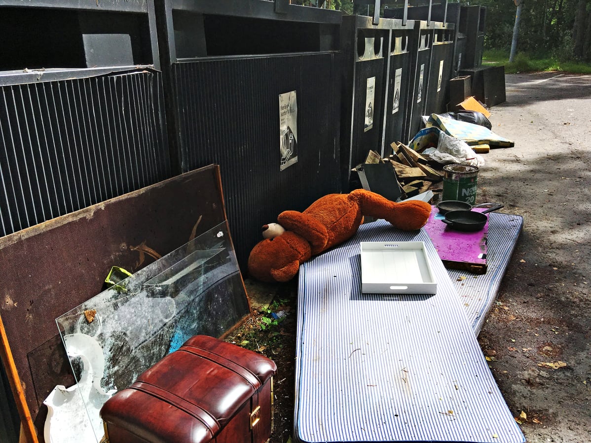 Kierrätyskartonki-keräysastian edessä lojuu patjoja, paistinpannuja ja pehmonalle. Samoin muidenkin keräyspisteiden edessä on sekalaista rojua.