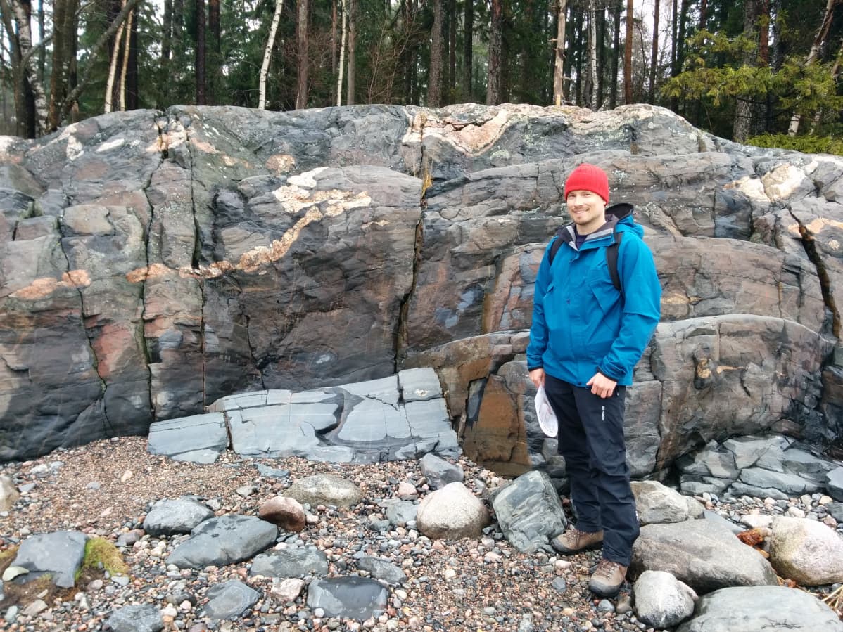 Itä-Helsingissä sijaitsevan Uutelan kosteaa rantakalliota, jonka juuressa pyöristyneitä rantakiviä. Kallion edessä seisoo punainen pipo päässään akatemiantutkija Jussi Heinonen.