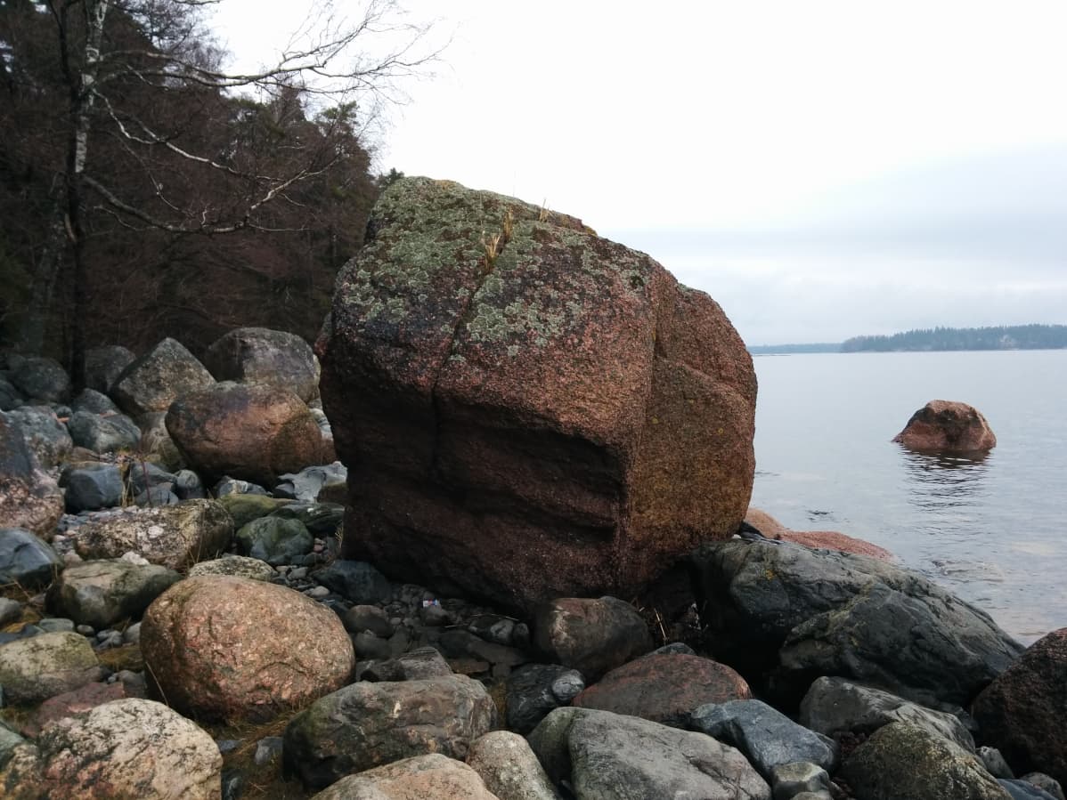 Kaksi metriä korkea punertava kivenlohkare, jonka ympärillä pienempiä muita kiviä. Taustalla näkyy melkein tyyni meri.