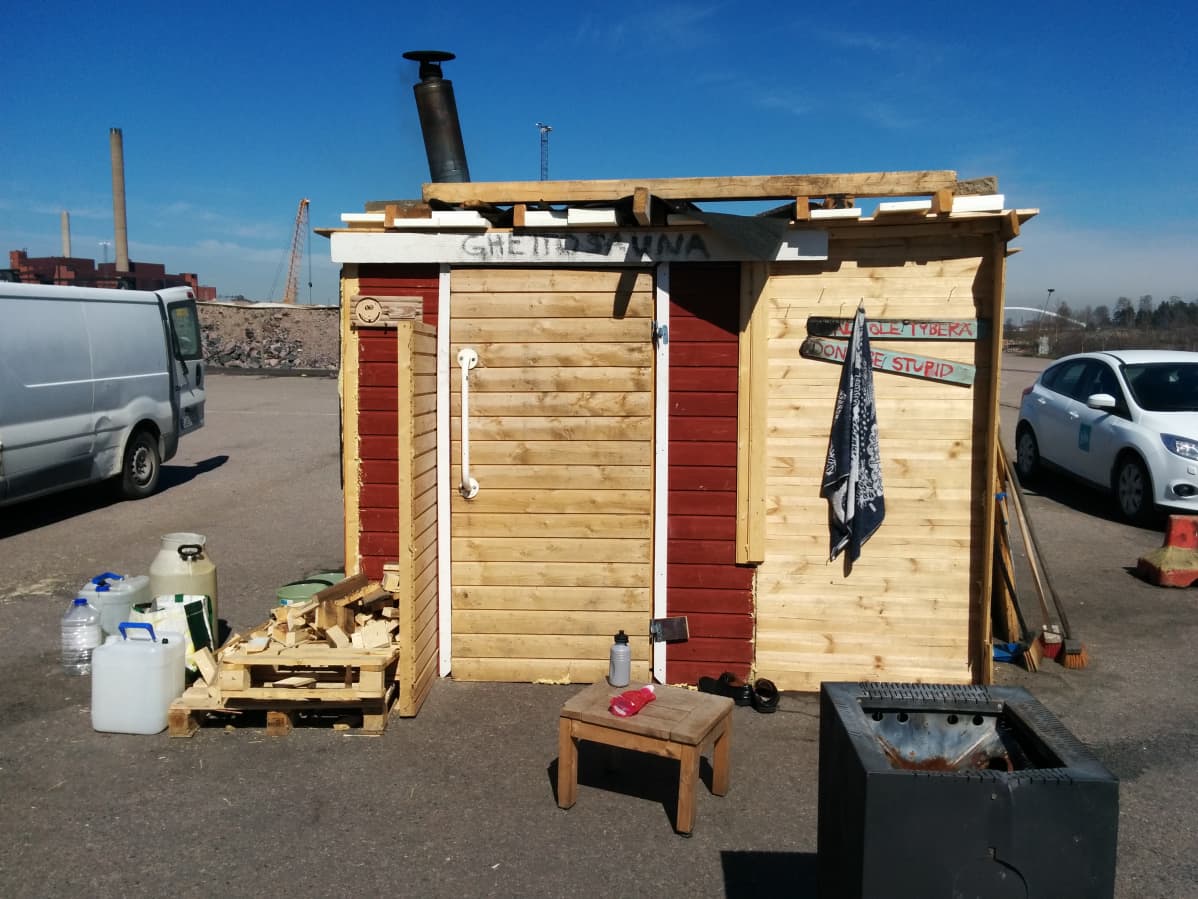 Hökkelin näköinen, lautarakenteinen väliaikainen sauna. Rakennuksen edessä vesiastioita ja polttopuita. Seinässä lukee "ghettosauna" ja "Älä ole typerä".