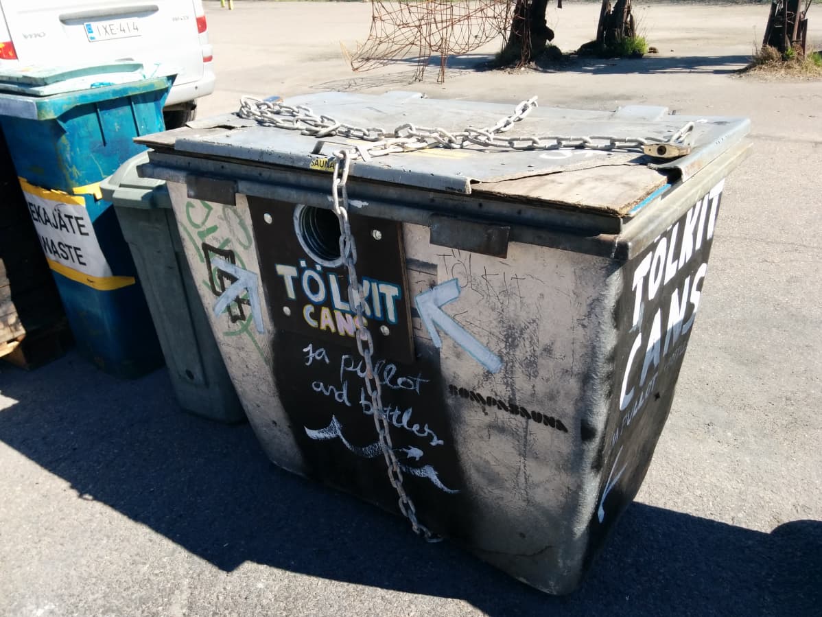 Harmaa roskalaatikko, jonka ympäri on sidottu kettingit. Laatikon kyljessä lukee tölkit.
