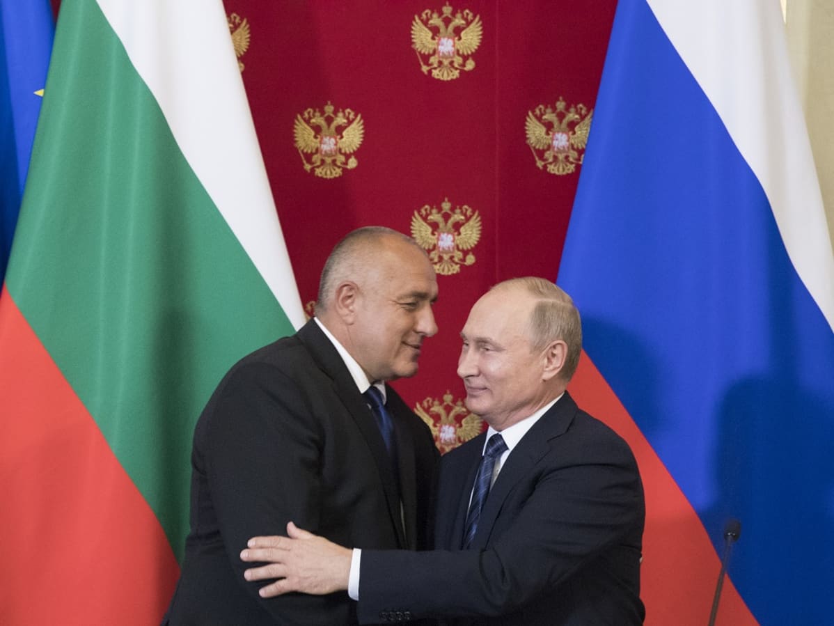 Borisov ja Putin syleilevät. Taustalla ovat Bulgarian ja Venäjän liput ja Venäjän kaksipäistä vaakunakotkaa esittävillä kuvilla koristeltu punainen kangas.