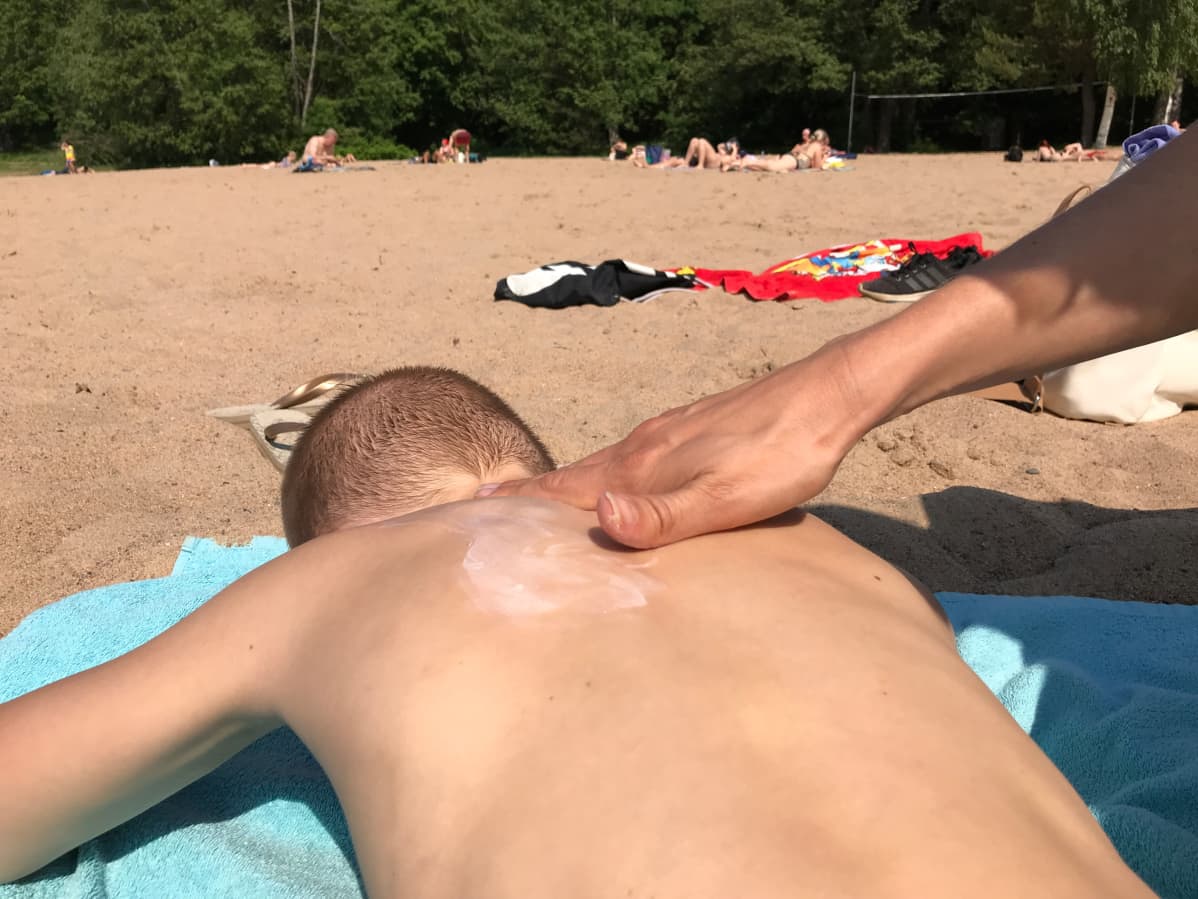 Käsi levittää aurinkorasvaa lapsen selkään uimarannalla.