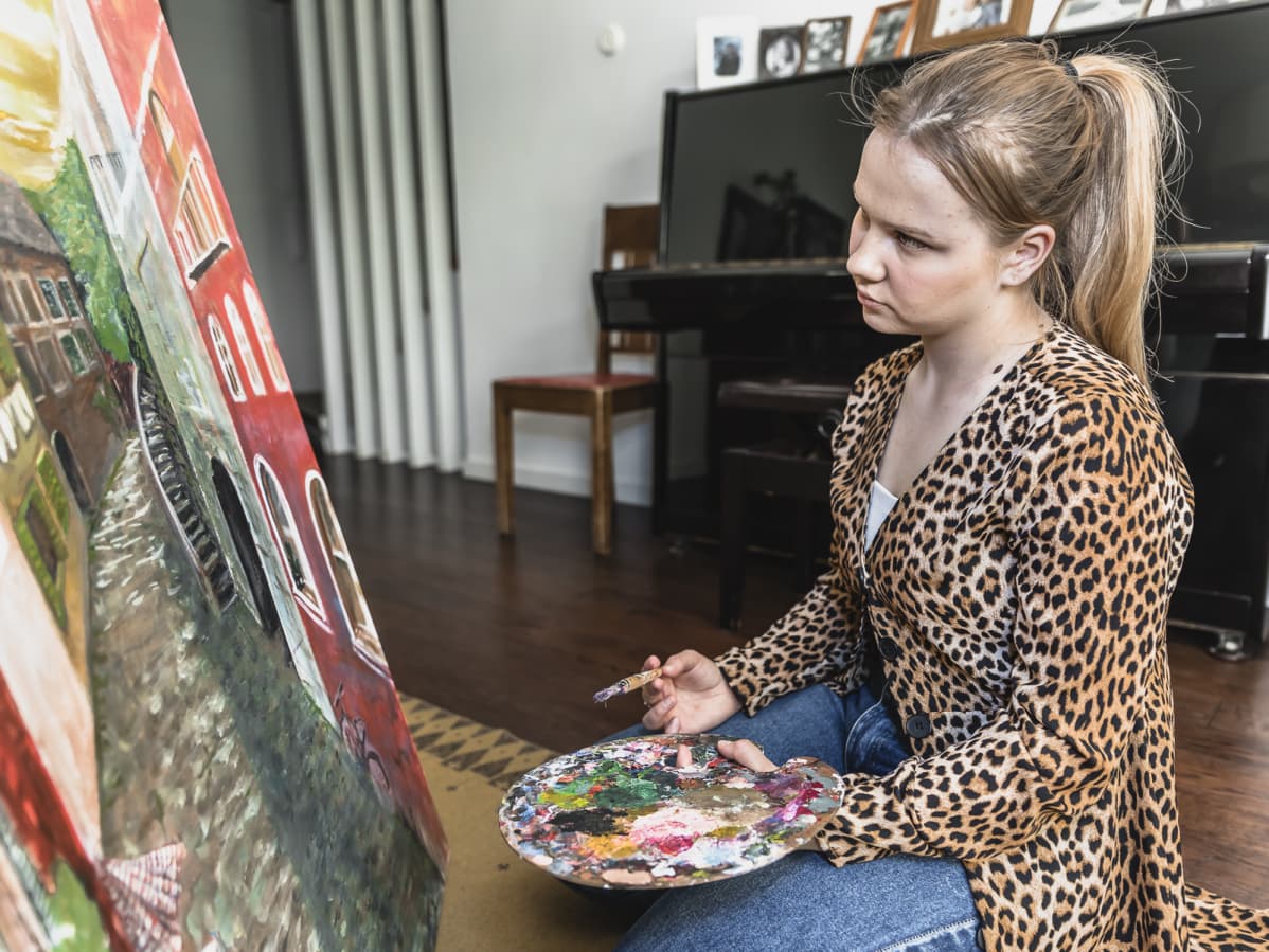 Amalia Paananen istuu polvillaan lattialla maalaustelineen edessä ja maalaa taulua.l