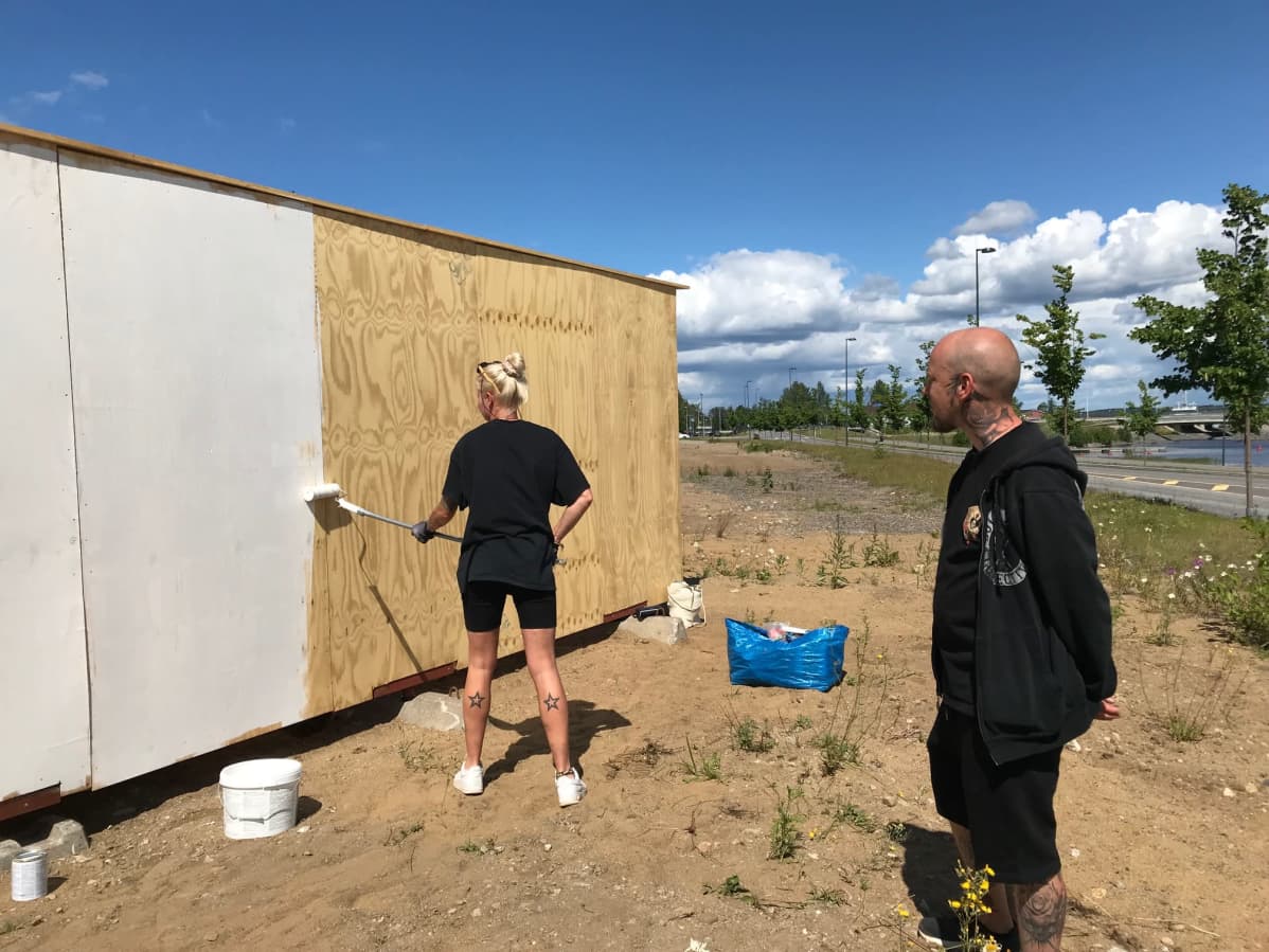 Inna Pulkkinen eli Poppamaija maalaa telalla vanerista graffitiseinää ja Toni Korhola katselee työskentelyä.