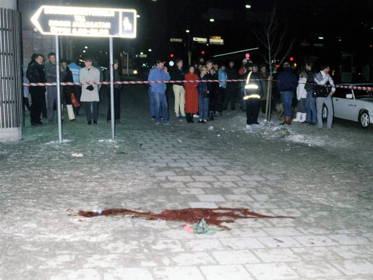 Kadulla on veriläikkä. Paikka on eristetty poliisin nauhalla. Eristyksen ulkopuolella seisoo ihmisiä.